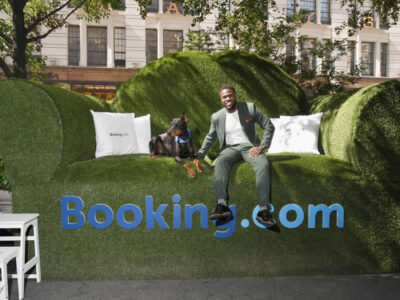 A man in a suit sitting in a large green chair with a dog, and the text 'Booking.com' below.